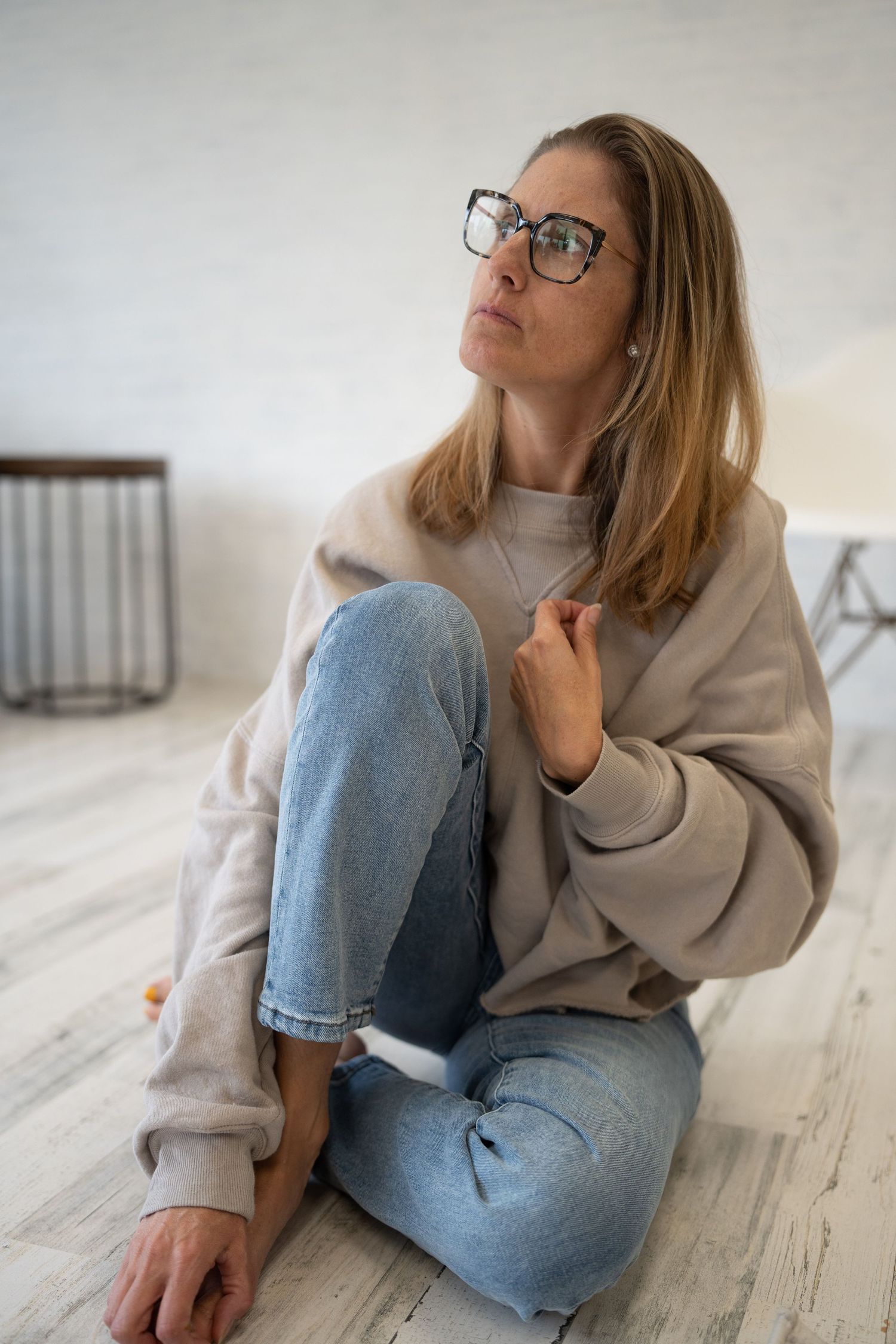 Casual portrait of person in oversized sweater and jeans sitting on light wooden floor.