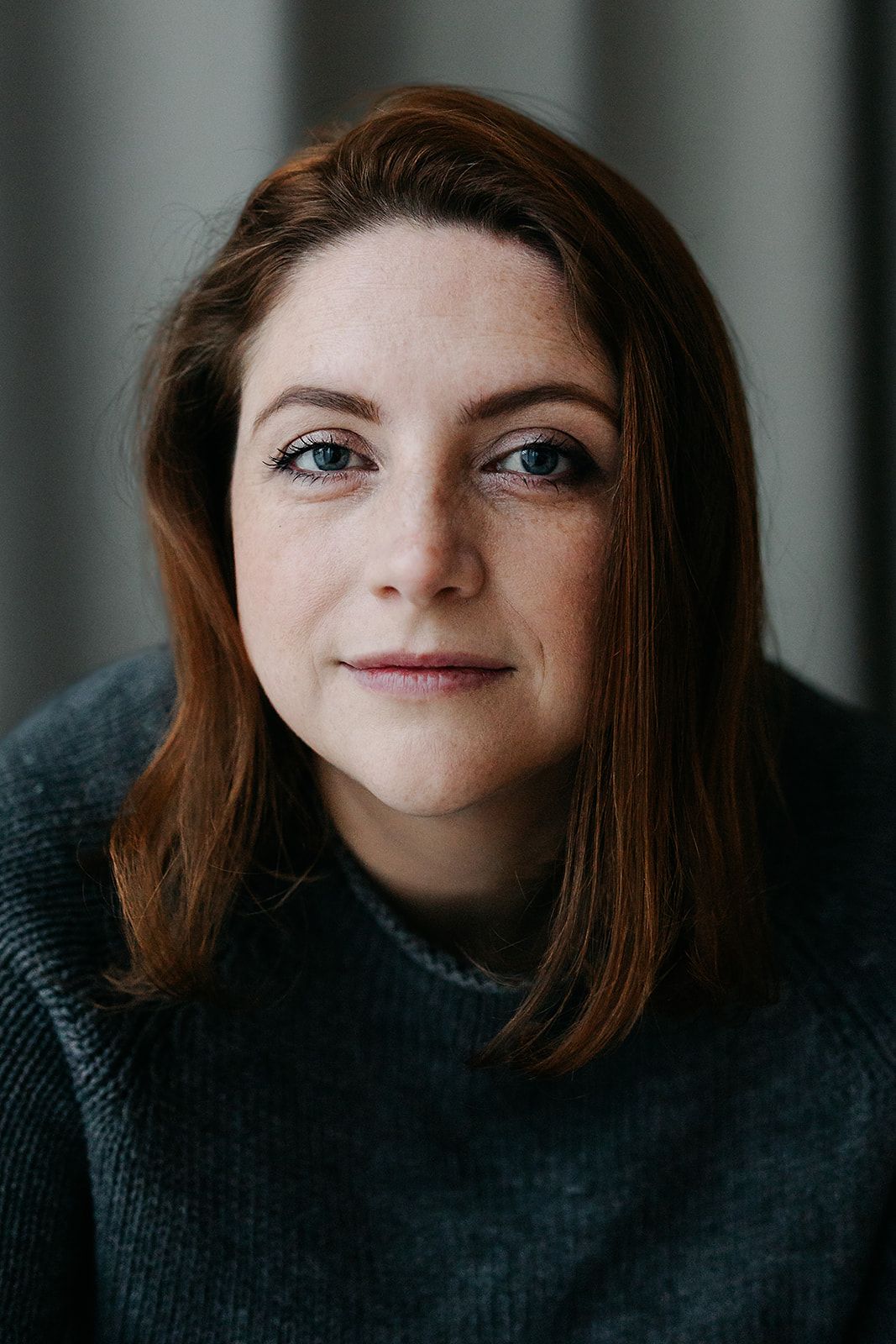 Portrait of someone wearing a dark sweater with soft lighting and natural makeup against a blurred background.