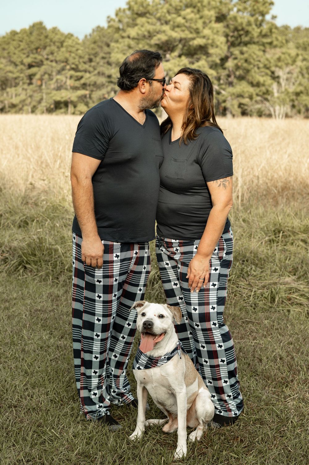 A couple in matching plaid pajama pants share romantic moments with their white dog in a golden field at sunset.