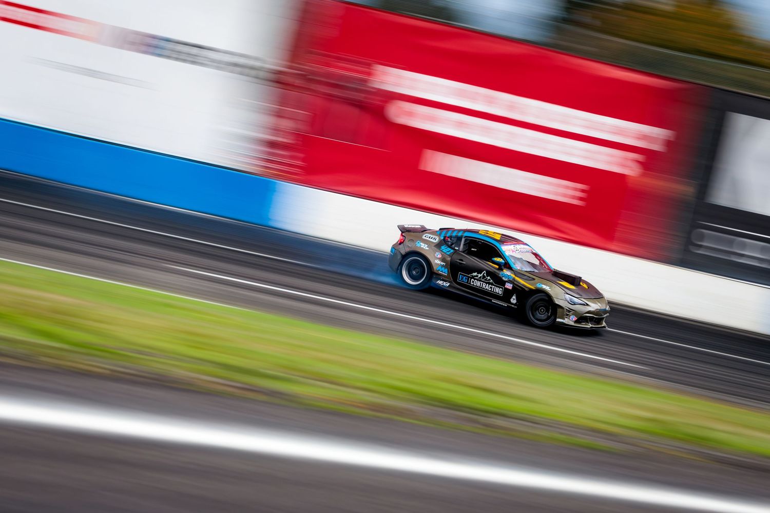 A red drift car slides sideways through a smoky turn during a professional drifting competition event.