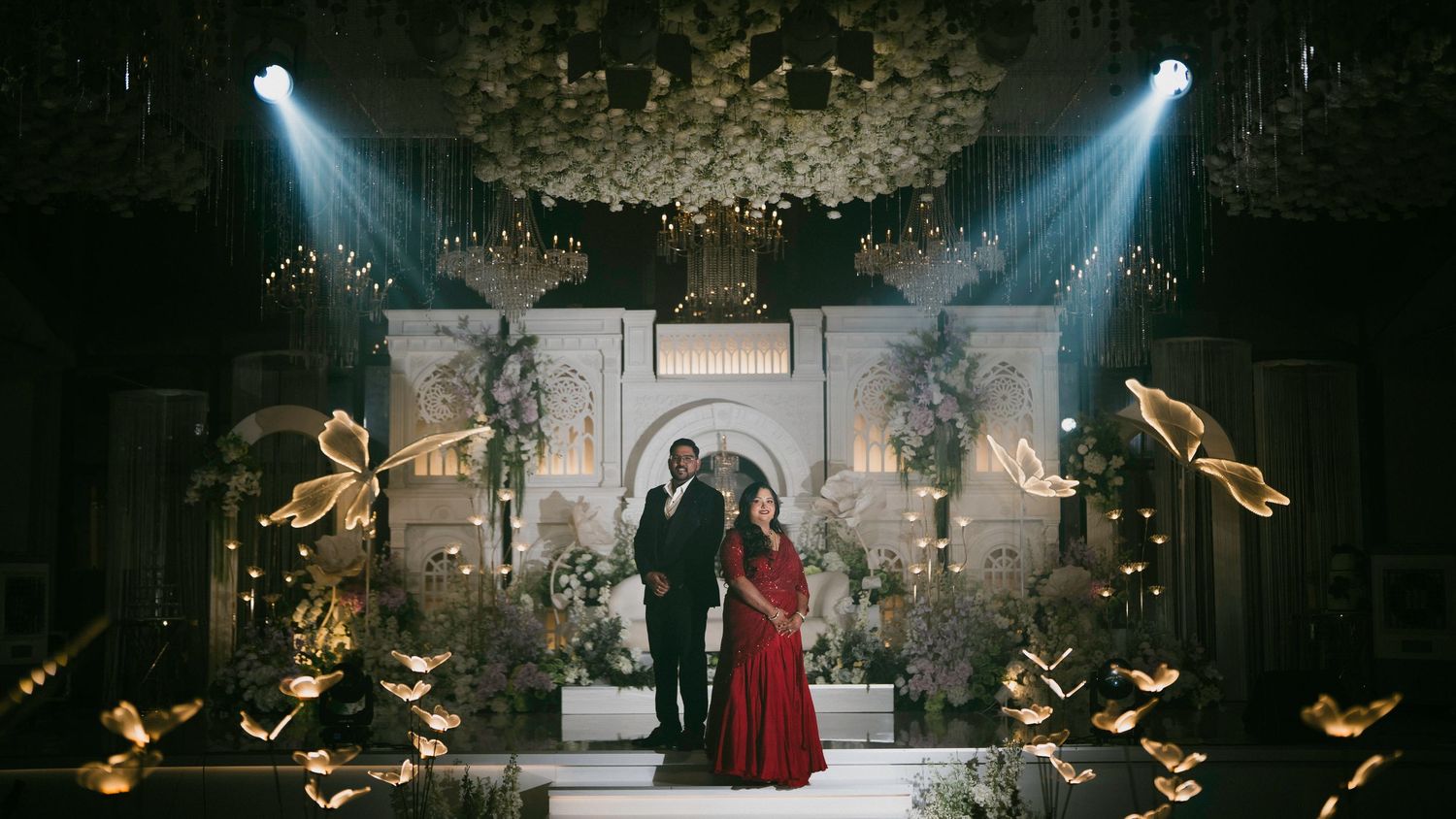 Couple posing at a grand wedding reception stage with golden butterfly decor and dramatic stage lighting.