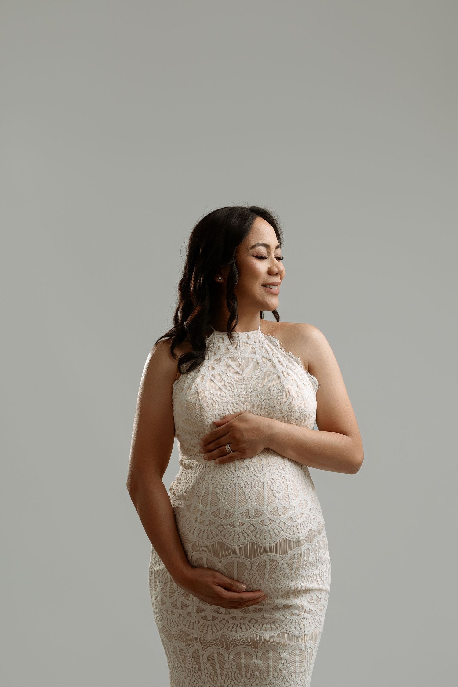 A woman in a white lace maternity dress poses in a series of photos showing her growing baby bump against a gray backdrop. Paloma Schell