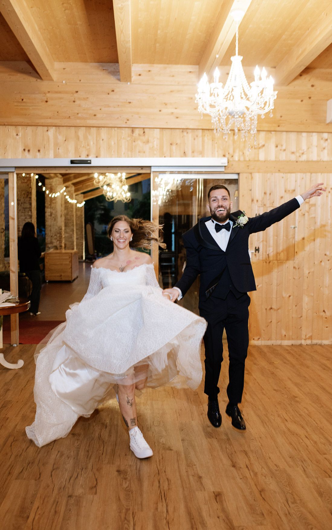 Bride and groom dance playfully across a wooden floor under elegant chandeliers in a rustic venue.