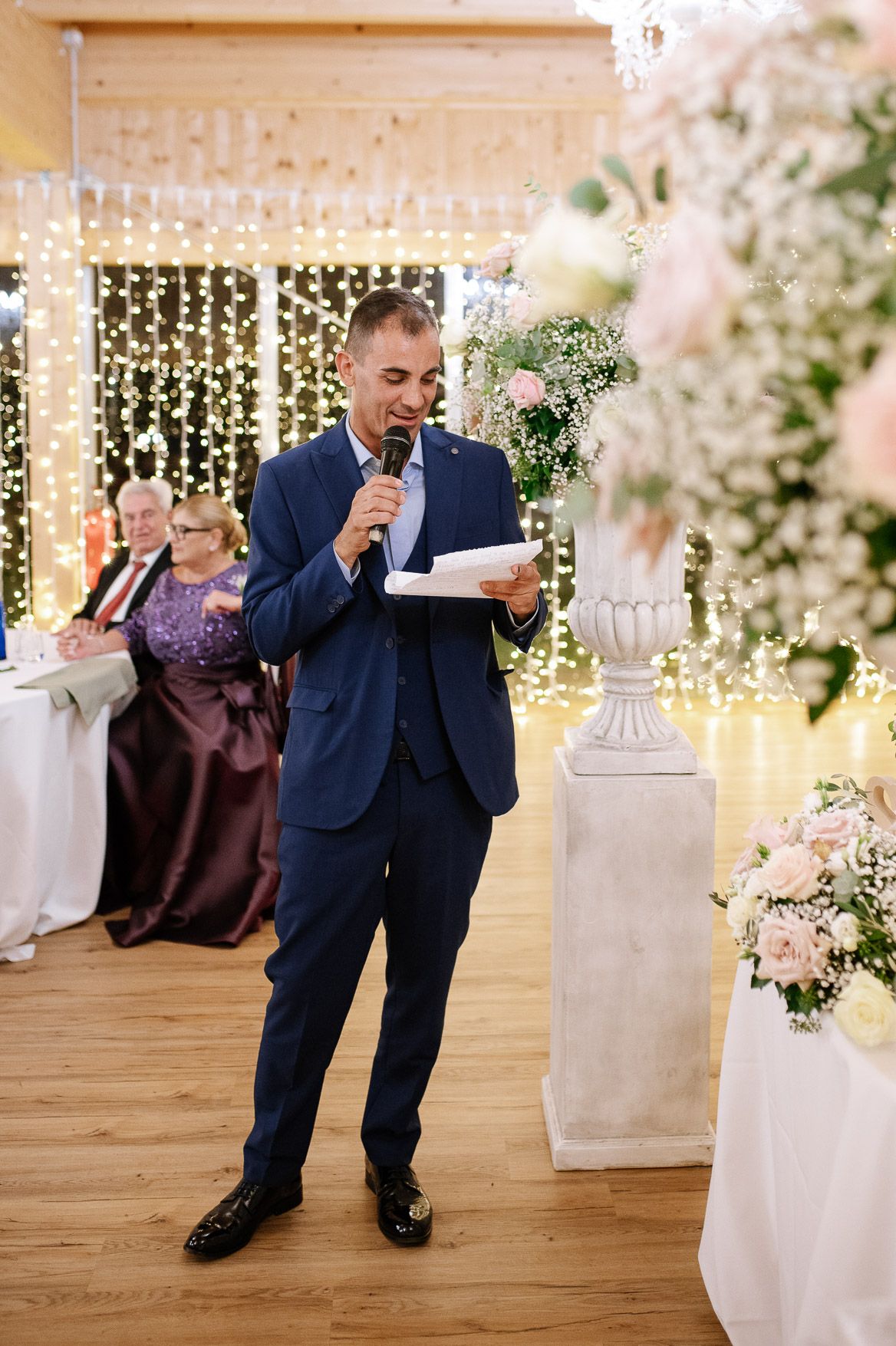 A speaker delivers remarks at an elegant wedding reception decorated with string lights and white floral arrangements.