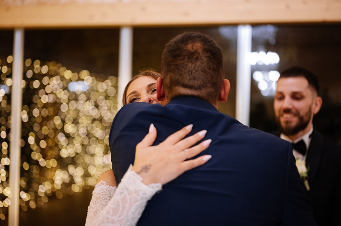 Two people embrace in an emotional hug at a wedding reception with glowing string lights in the background.