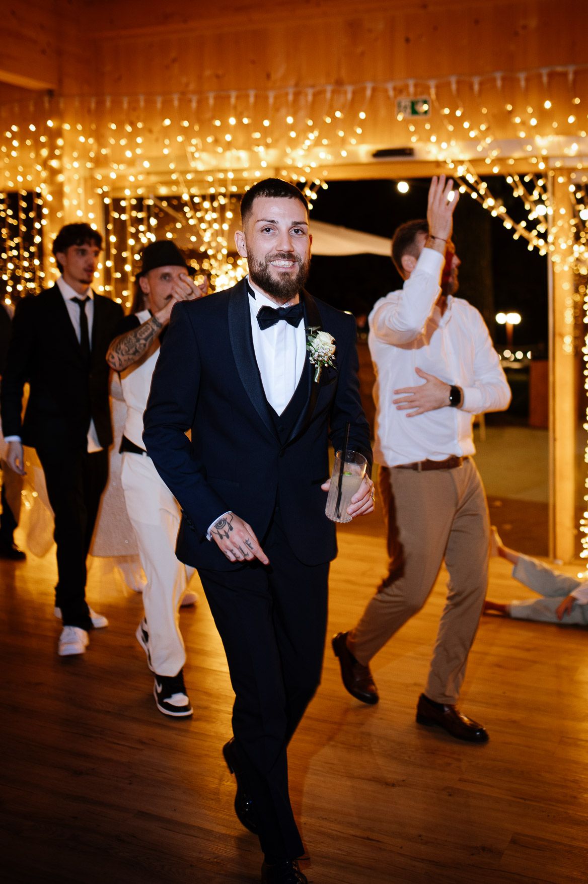 Wedding reception guests dance on a wooden floor illuminated by warm string lights overhead during evening celebrations.