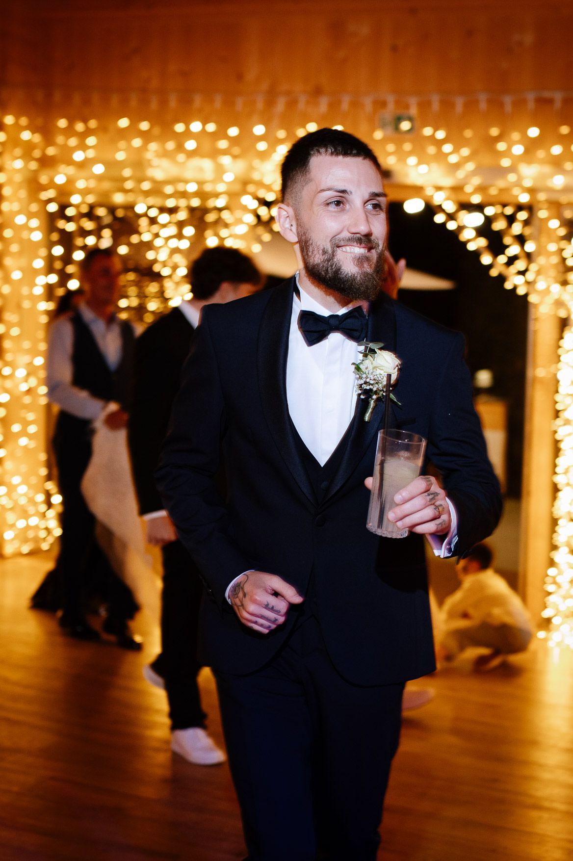 Groomsman in tuxedo holding drinks at wedding reception with twinkle light backdrop.