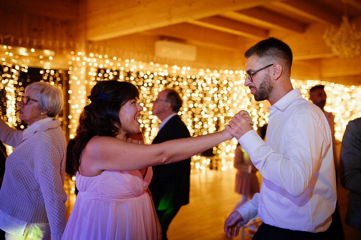 People dancing and celebrating at a wedding reception with glowing string lights decorating the walls in warm ambient lighting.