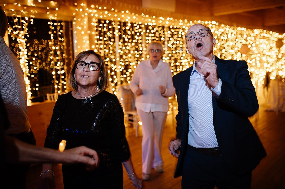 Wedding reception guests dancing and singing enthusiastically under glowing string lights.