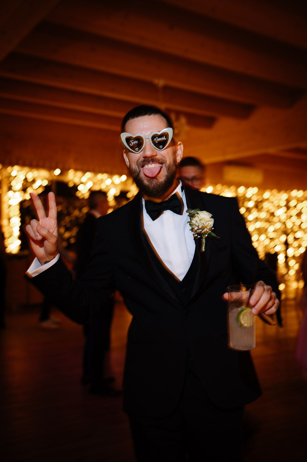 Wedding guests in formal black attire dance and celebrate under warm string lights at an indoor reception.