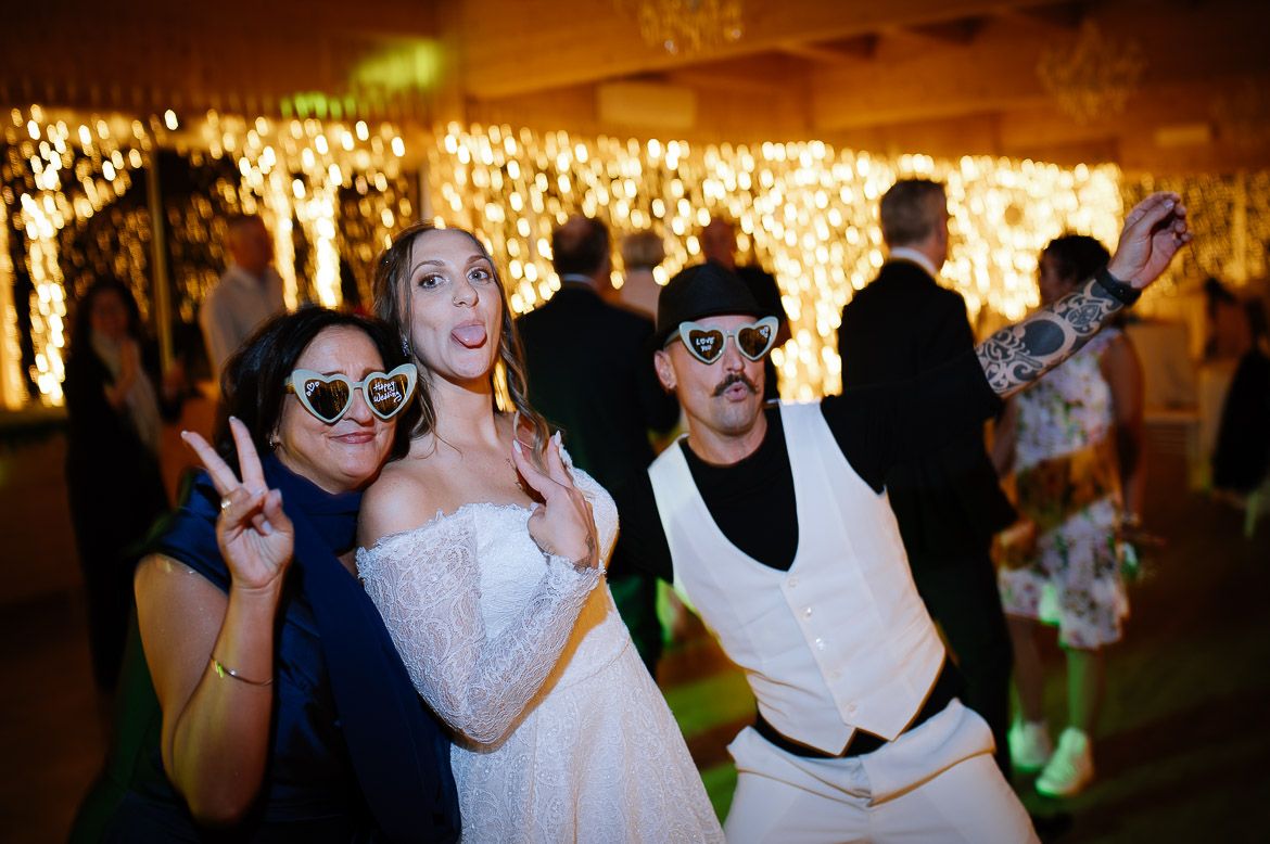 Wedding reception party scene with guests dancing and celebrating in a warmly lit indoor venue.