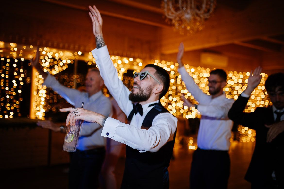 Couples dancing at a rustic wedding venue illuminated by warm string lights and chandeliers.