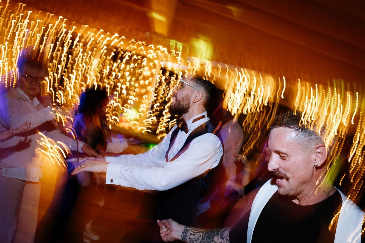 Wedding guests dancing and celebrating on a dance floor under warm glowing lights at an evening reception.