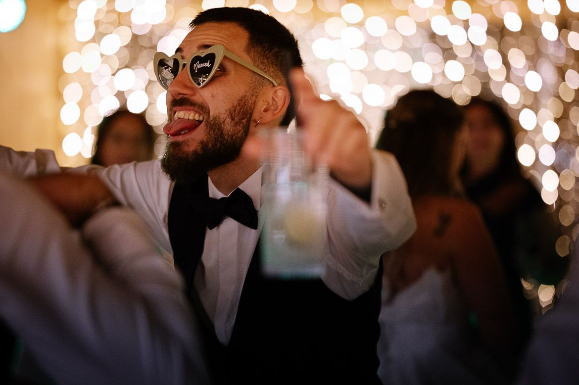 Wedding guests dance and celebrate on a dimly lit dance floor with glowing bokeh lights in the background.