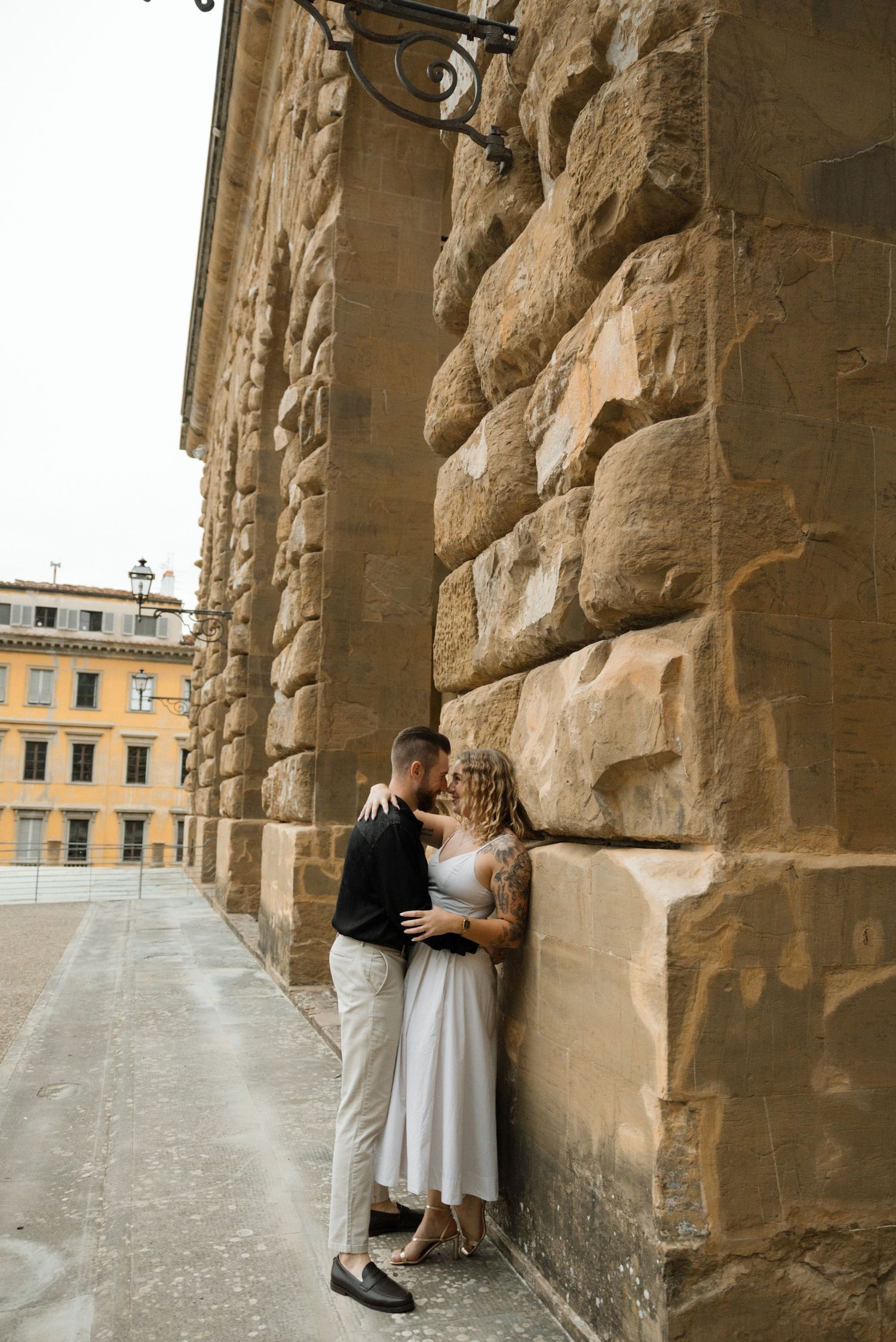 Romantic moment captured under historic stone arches with yellow buildings visible in background.
