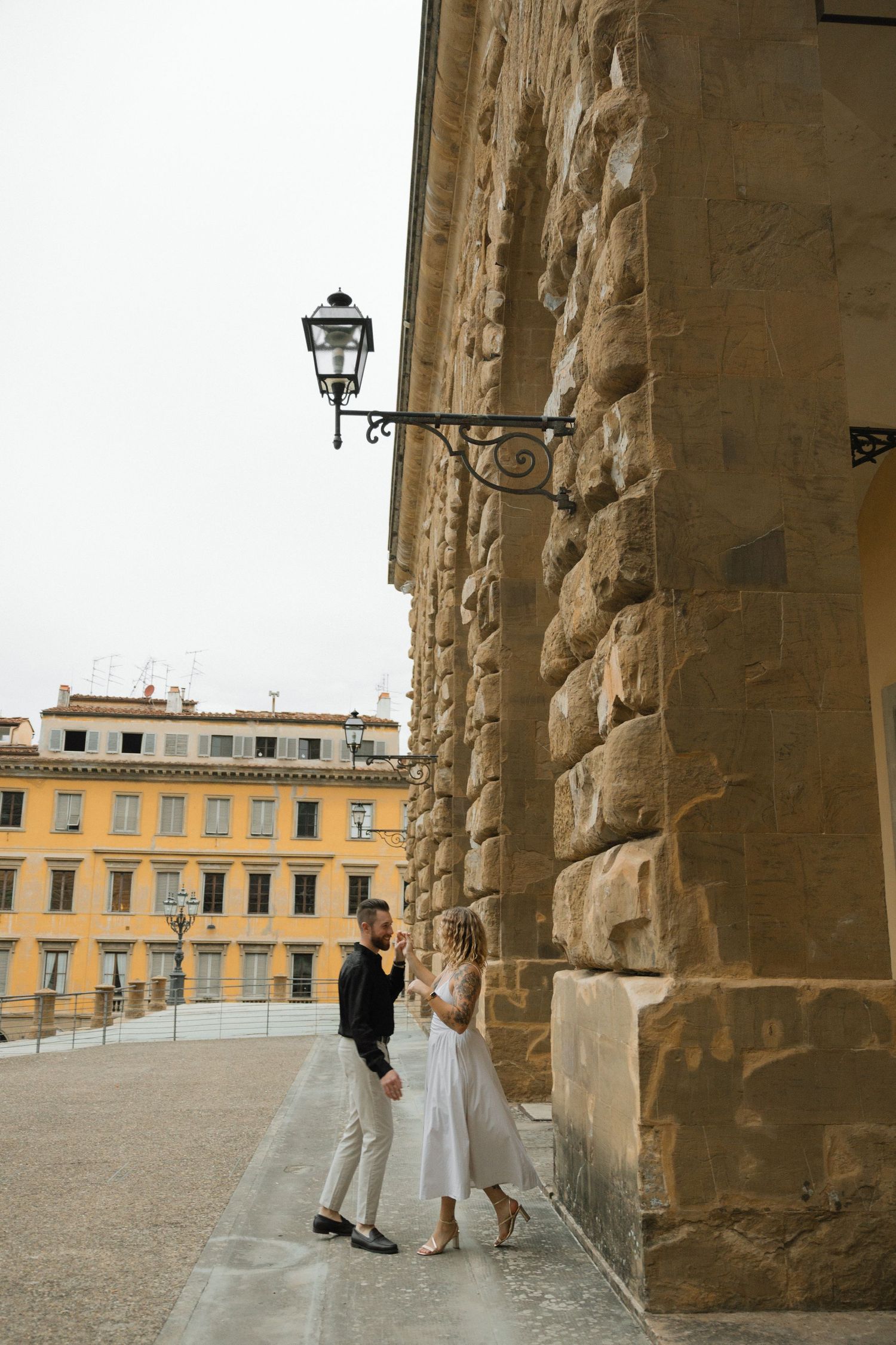 Couple posing along a historic stone colonnade with yellow Italian buildings in the distance.