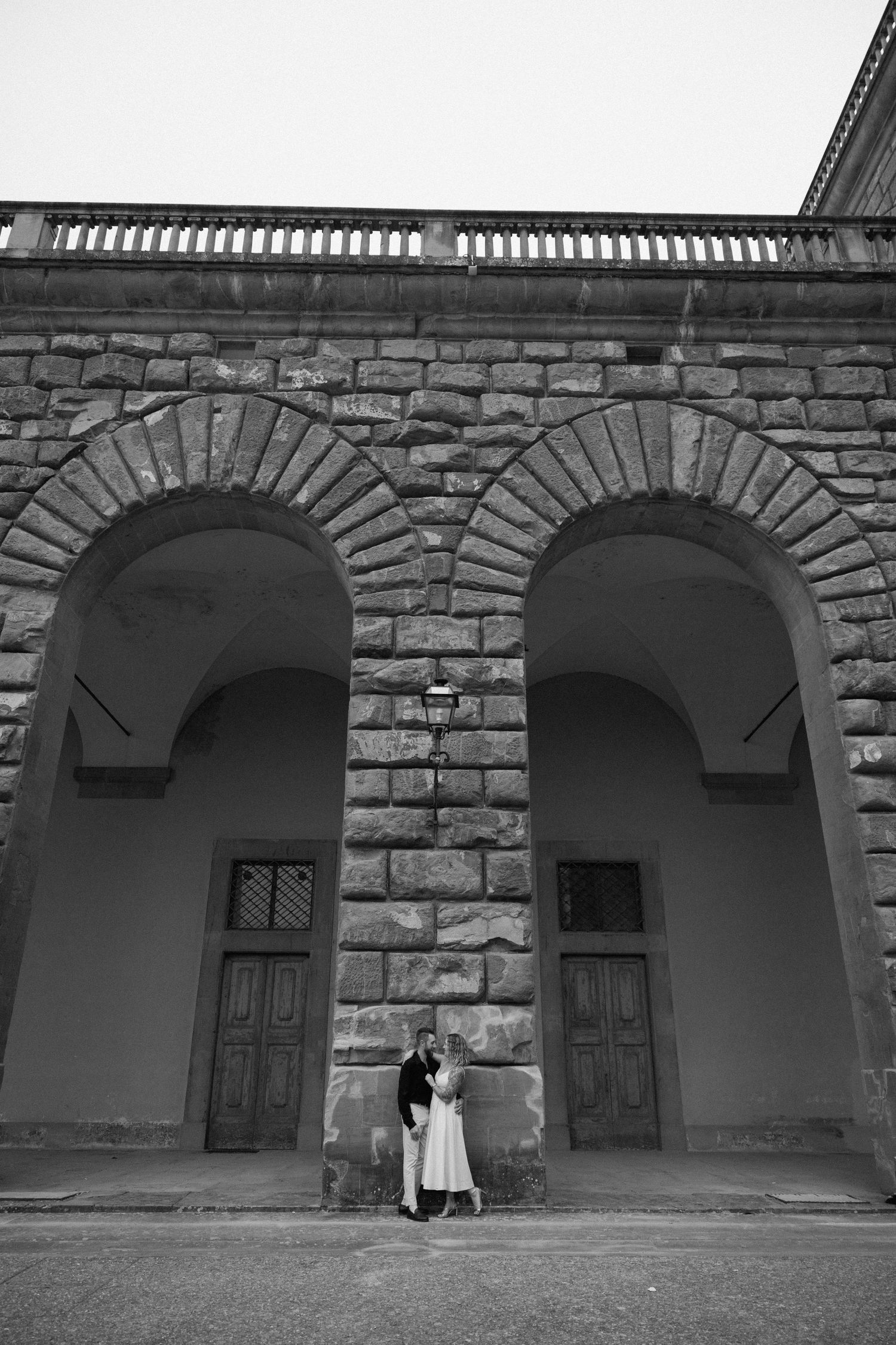 A series of stone archways with wooden doors along a historic building wall in black and white.