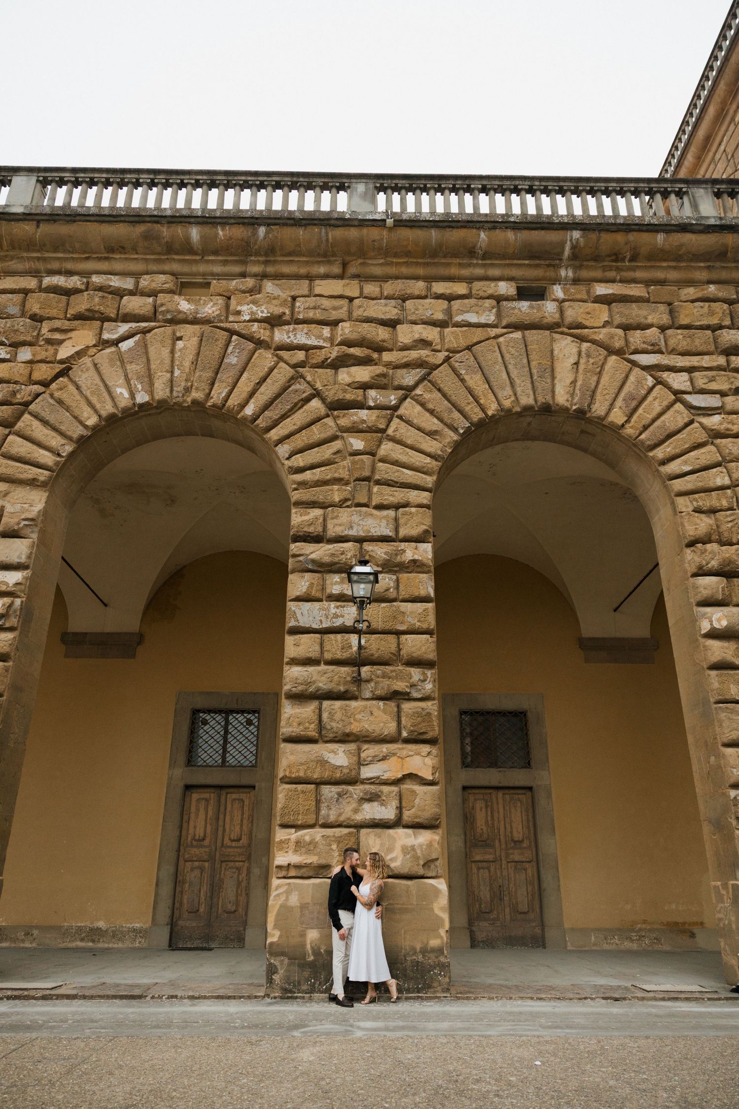A couple stands beneath grand stone archways of a historic Italian building.
