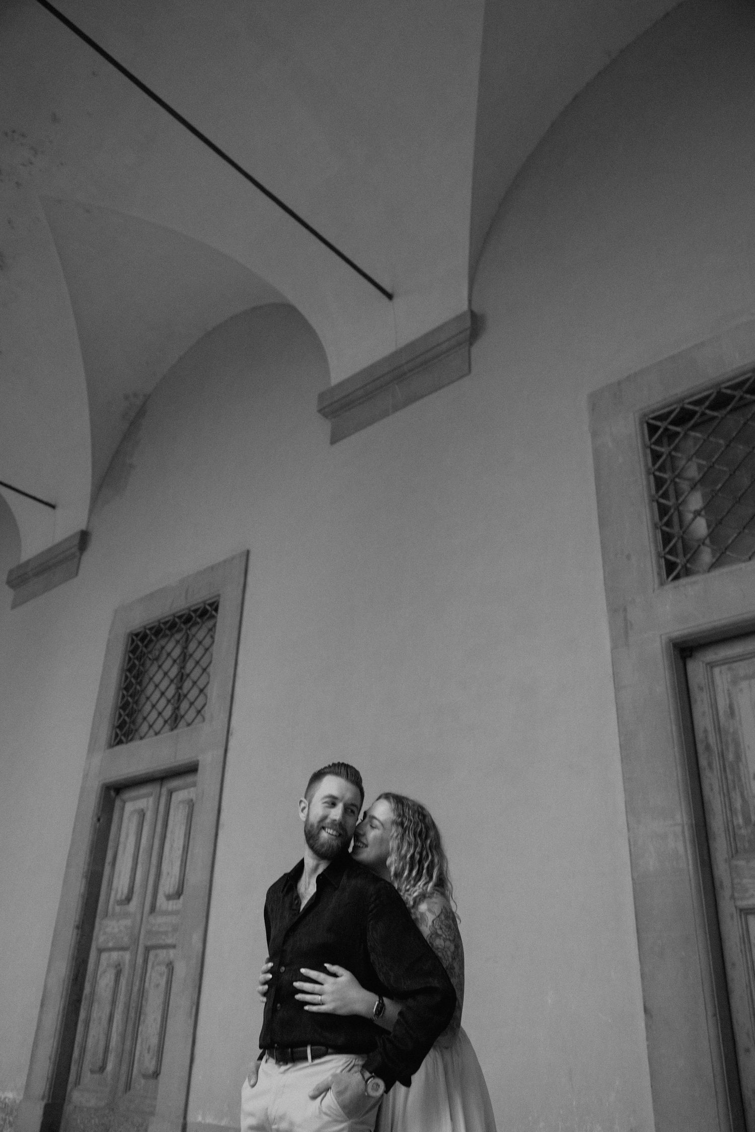 A romantic black and white portrait of a couple embracing under arched architecture with vintage doors.