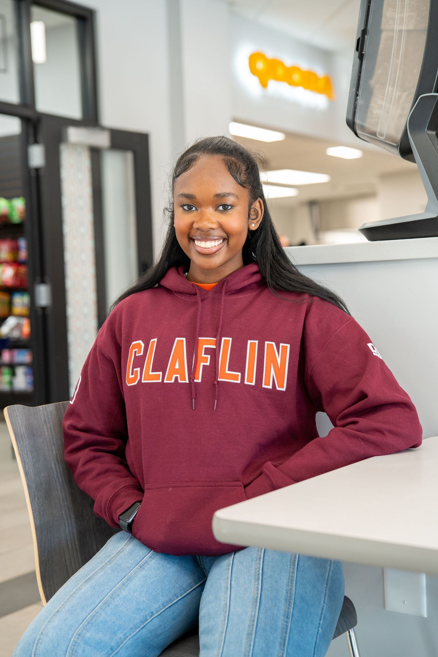 A smiling student in a maroon Claflin hoodie sits at a table in a campus dining area.