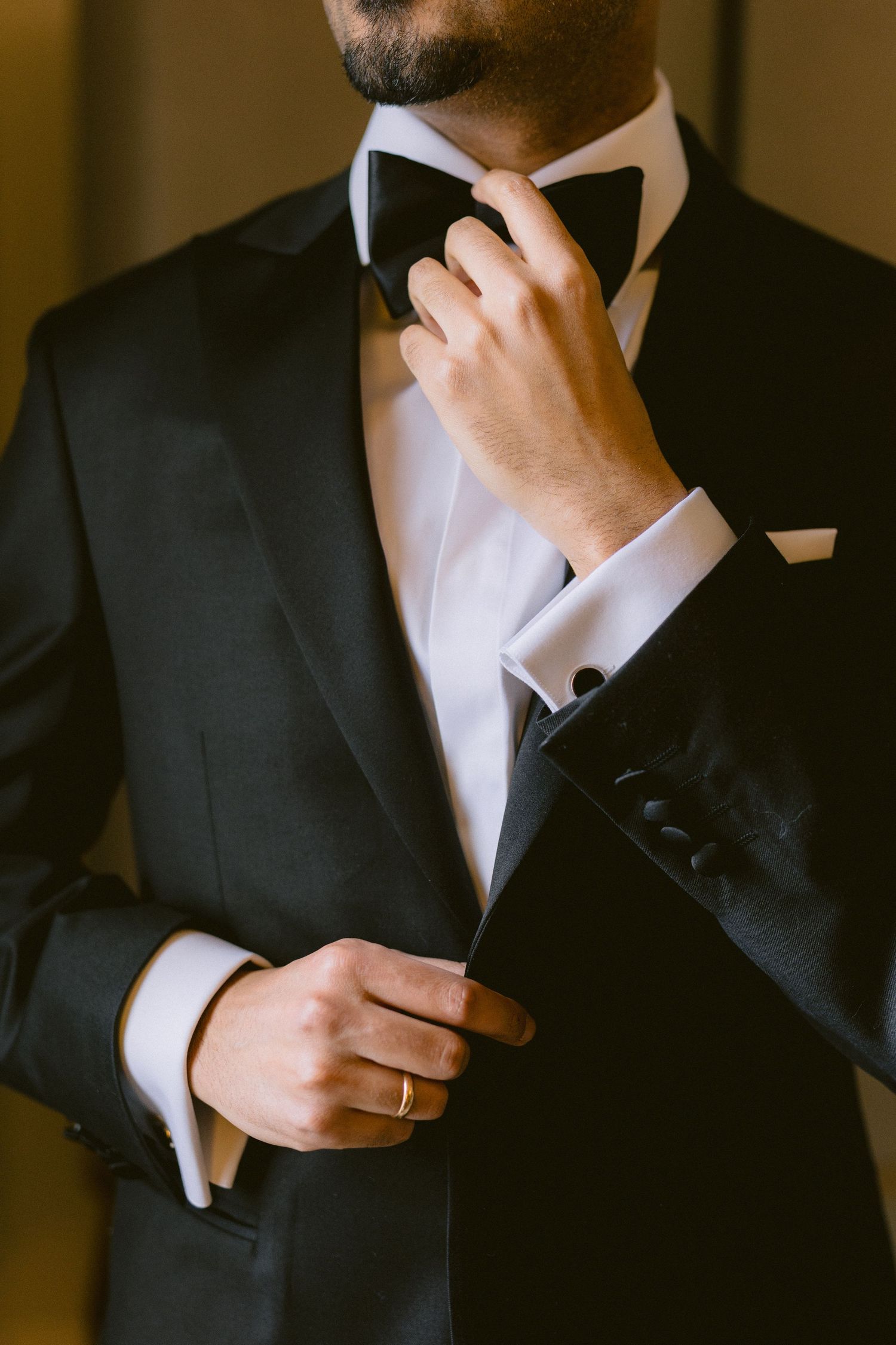 Close-up view of someone adjusting a black bow tie while wearing a formal tuxedo in warm lighting.