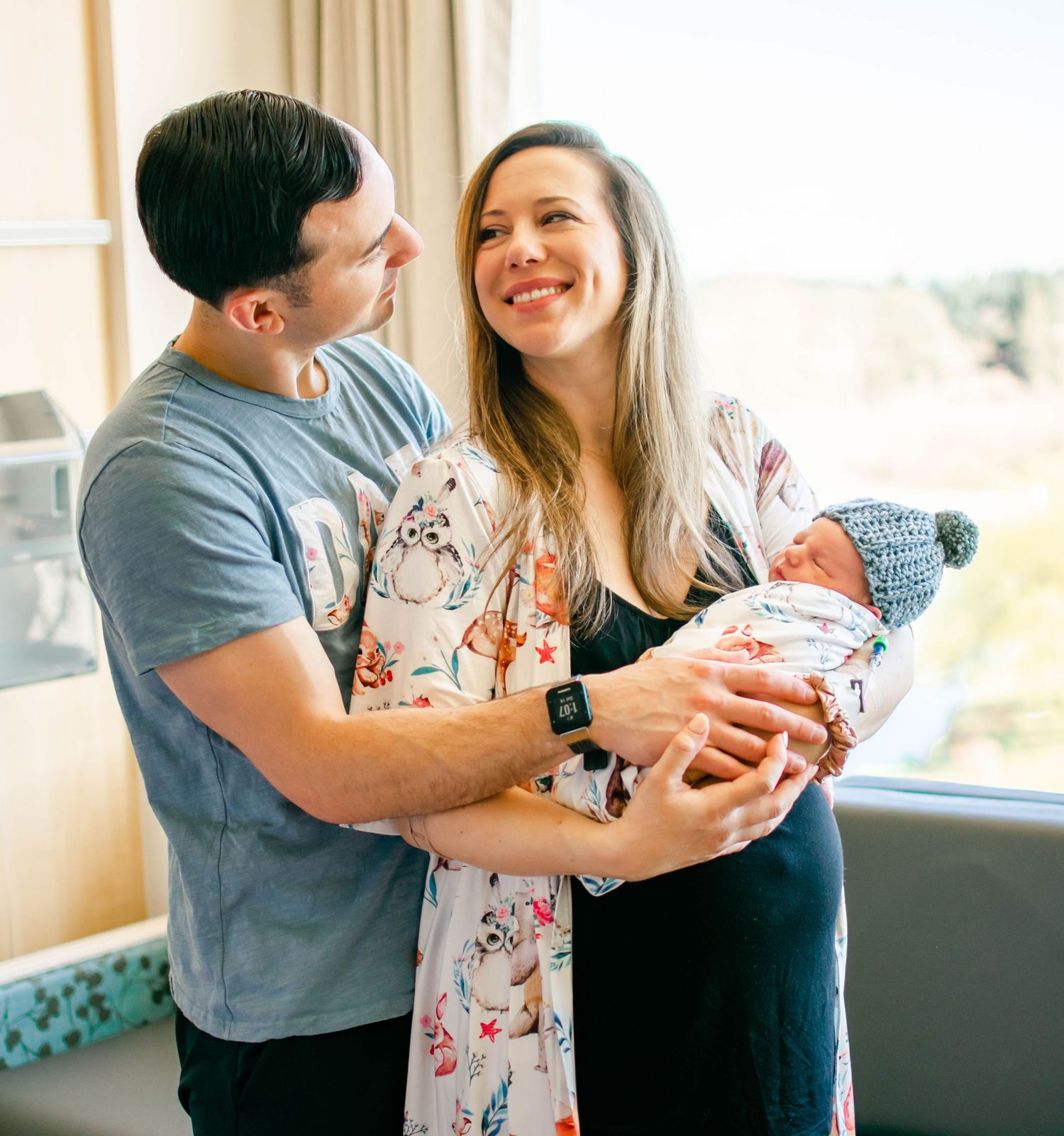 A couple stands by a window sharing a tender moment while holding a newborn baby wrapped in a blanket.