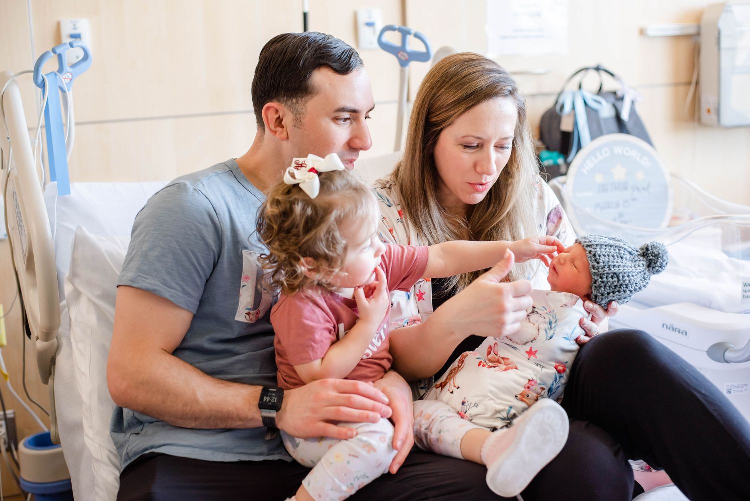 Family sitting together on a hospital bed with a newborn baby in a bright, cheerful room.