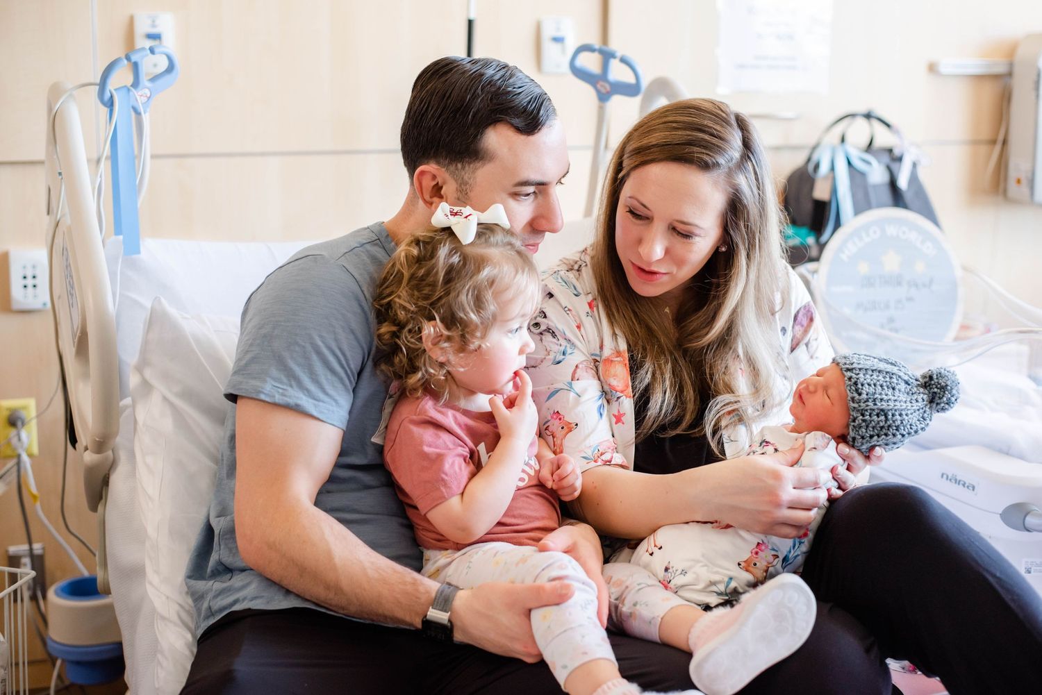 Family sits together on hospital bed with newborn baby and toddler in tender moment.