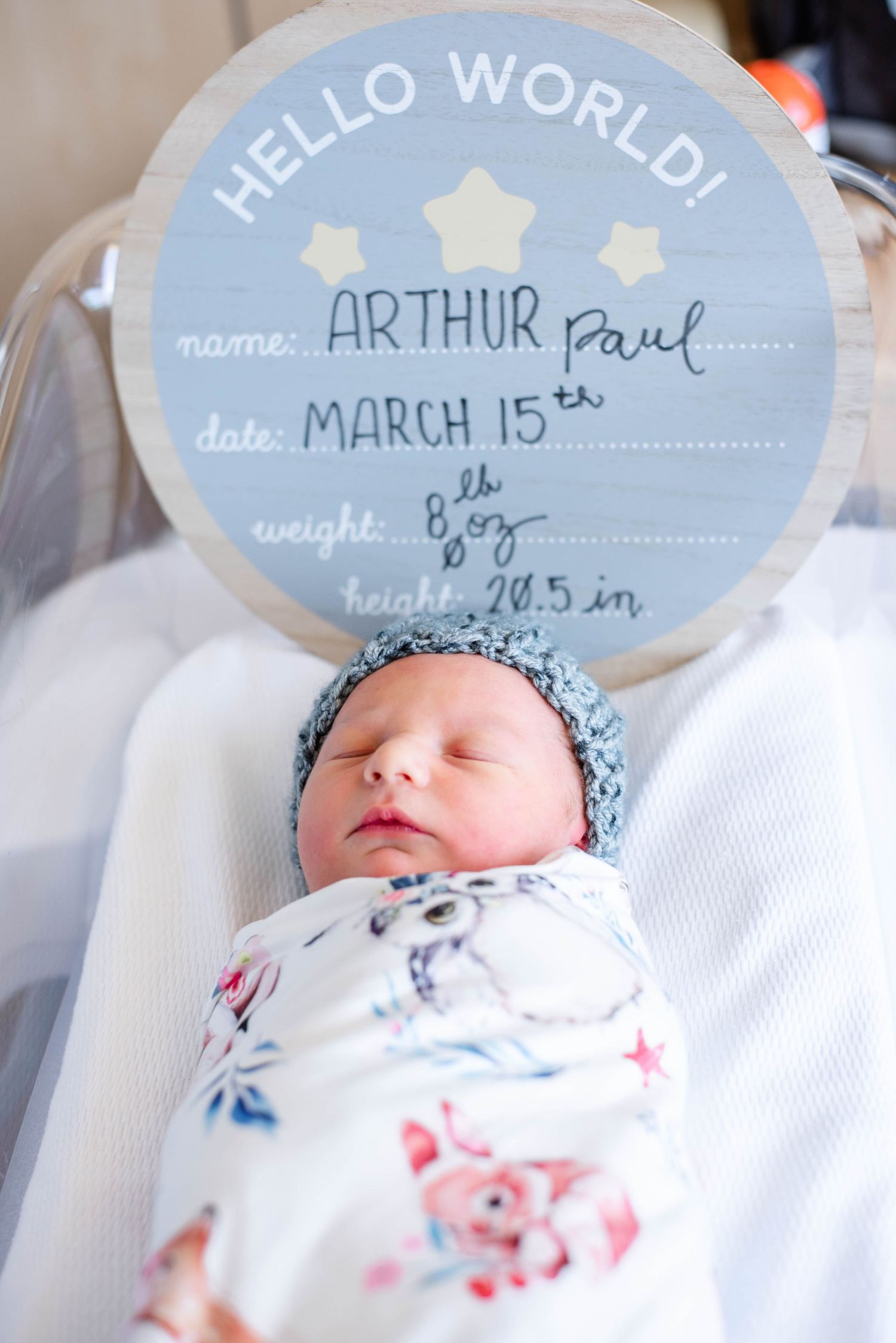 A sleeping newborn baby wrapped in floral swaddle next to a blue 'Hello World' announcement sign with birth details.