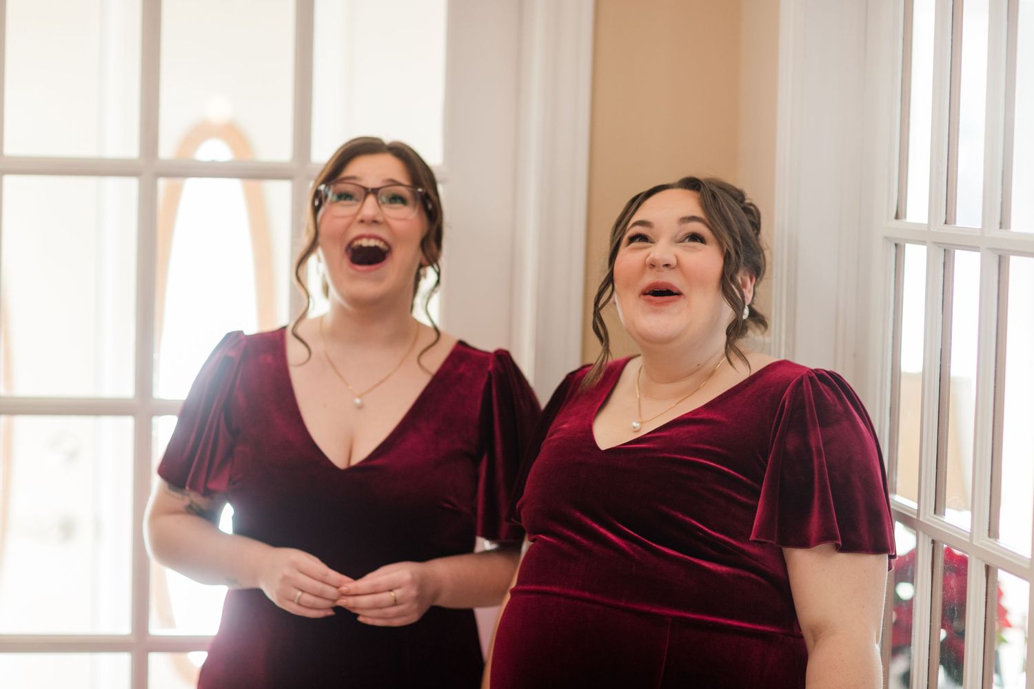 A group of people in burgundy dresses laugh together during a joyful moment in a bright indoor setting.