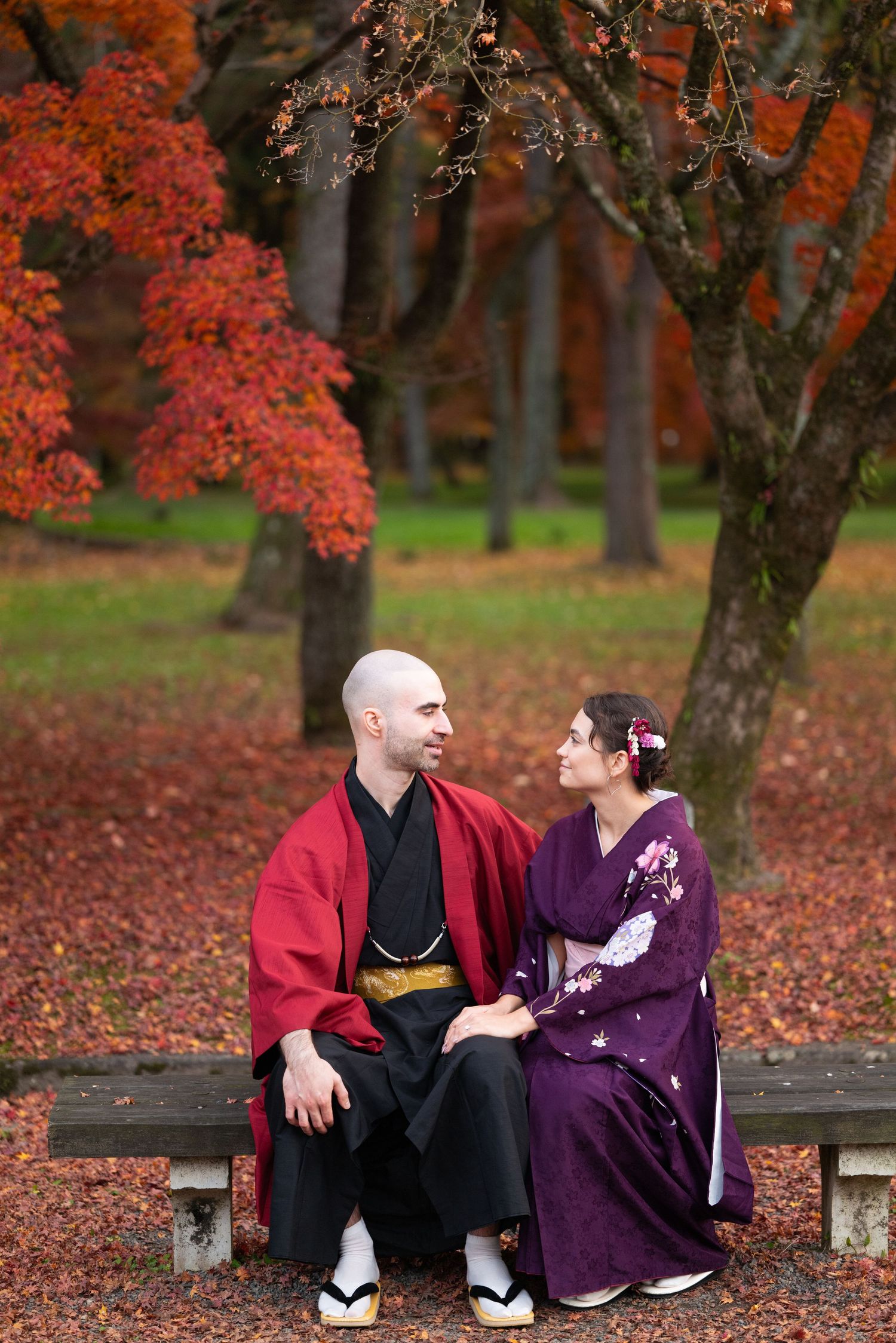 Couple dressed in red and purple kimono gazing at each other on a bench amid stunning fall foliage in Japan.