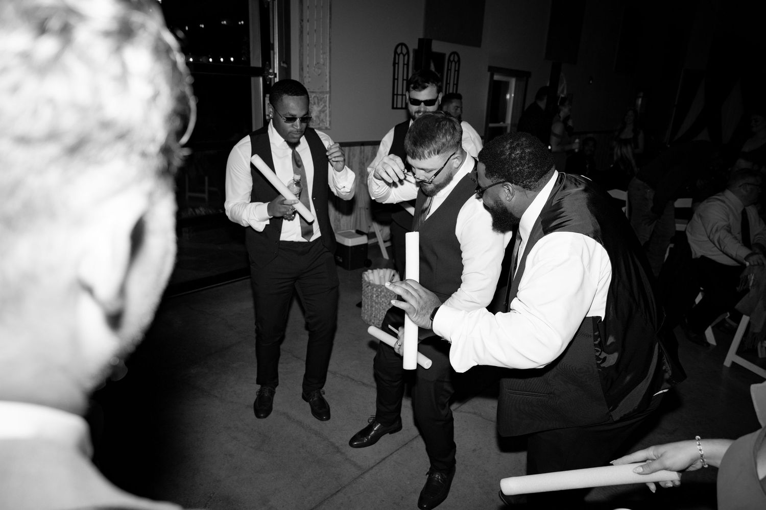 Black and white photo of people dancing at a lively party or reception in a dimly lit venue.