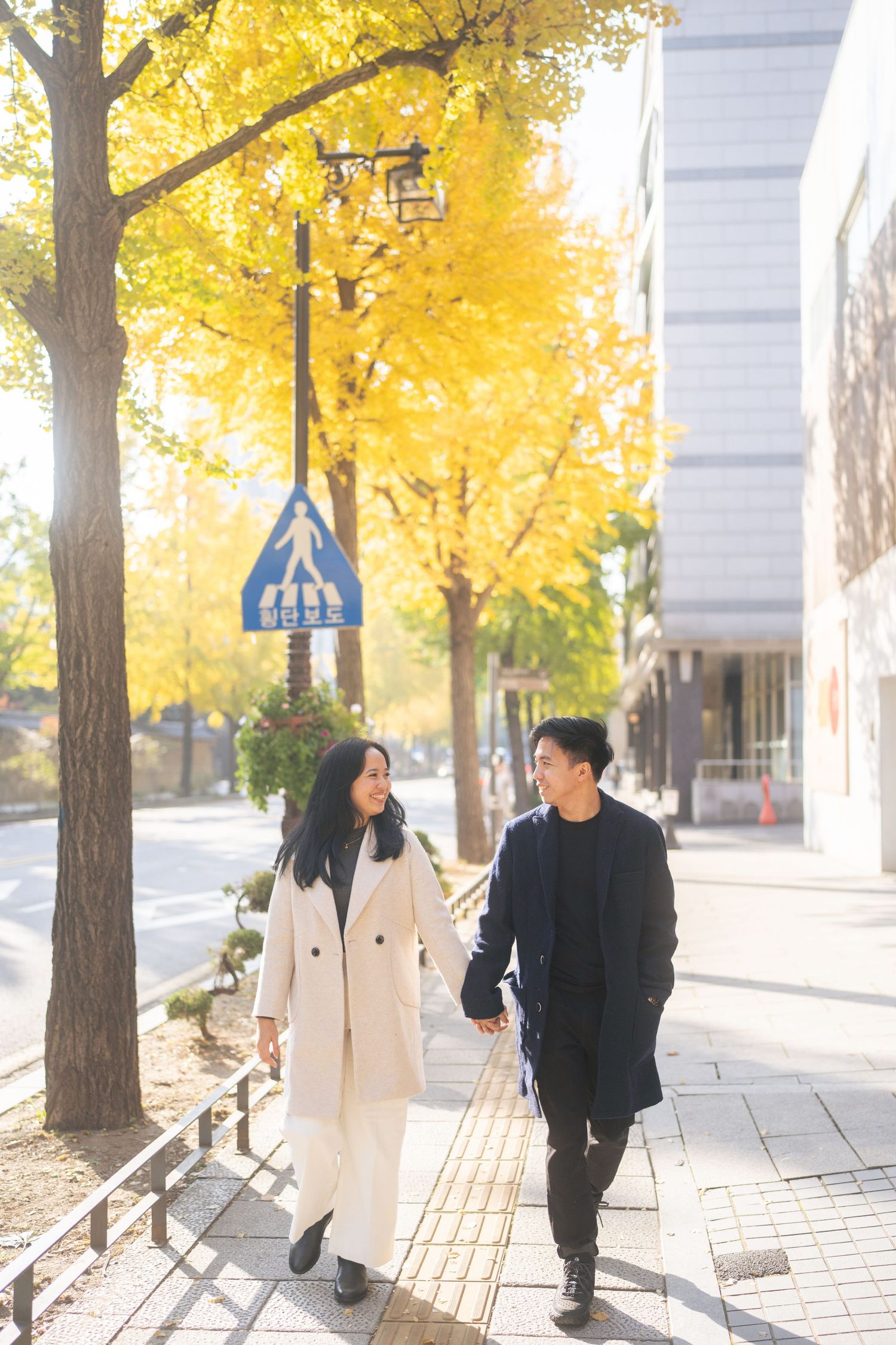 A couple walks together on a city sidewalk lined with golden autumn trees on a sunny afternoon.