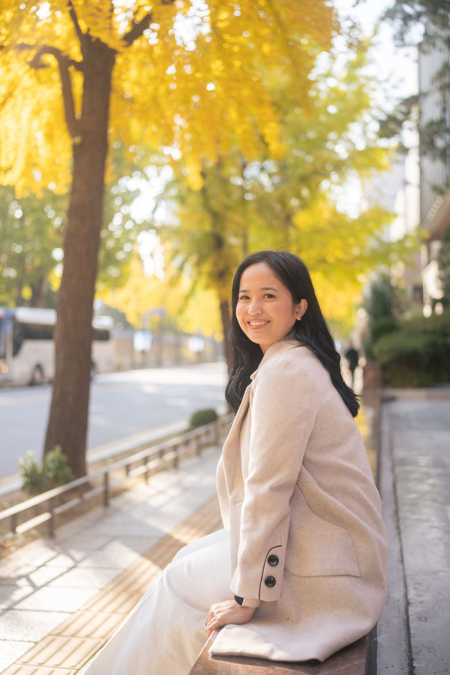 A person in a beige coat sits on a wooden bench under golden autumn trees on a sunny afternoon.