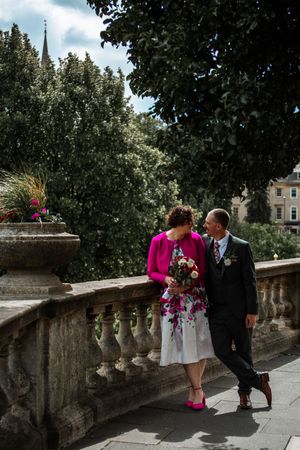 A couple stands along a stone balustrade with trees and blue sky in the background. Bath Guildhall Wedding