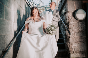 A bride in a flowing white wedding dress walks through an industrial-style hallway with exposed brick walls and pipes.