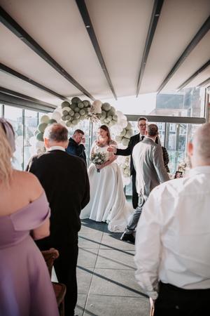 Wedding guests gather in a modern venue with white beams and large windows during a celebration.