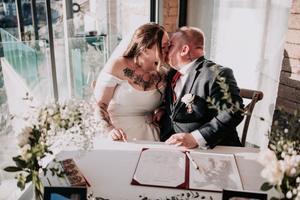 A romantic couple signs their wedding documents at a bright indoor venue surrounded by floral decorations.