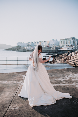 A flowing white wedding dress billows dramatically against a coastal backdrop with city buildings in the distance.