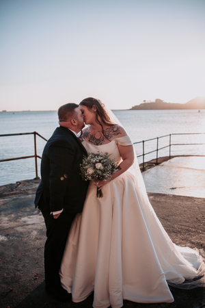 A romantic sunset wedding photo on a coastal overlook with the ocean and lighthouse in the background.