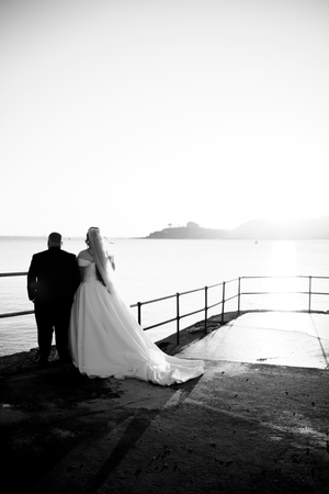 Silhouettes of a wedding couple standing by seaside railings at sunset create a romantic black and white scene.