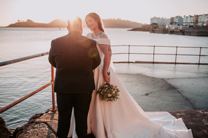 A romantic couple in wedding attire stands at the edge of a waterfront during golden hour.