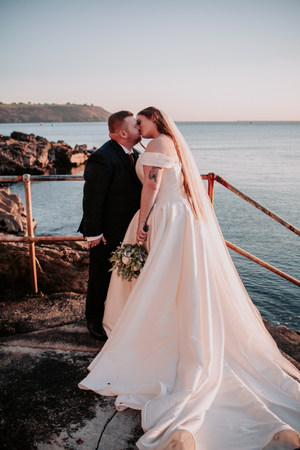 Couple in wedding attire share an intimate moment along a seaside railing at sunset.