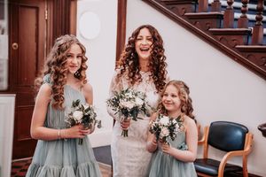 Three people in formal attire holding white and green floral bouquets stand together by a wooden staircase.