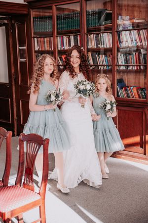 Bridesmaids in light blue dresses walk through a library holding white floral bouquets.