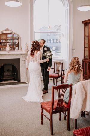A romantic first dance moment captured in a bright room with large windows and vintage wooden chairs.
