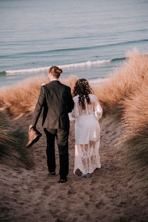 A newlywed couple walks hand in hand through beach grass along the shoreline at sunset.