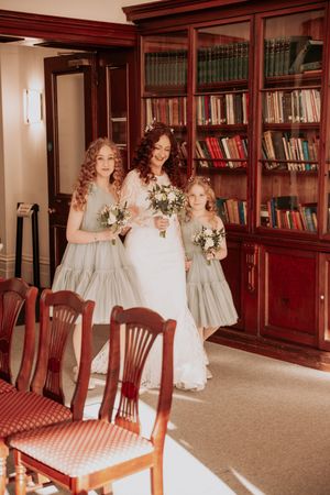 Wedding party members in light colored dresses gather in a library room with wooden bookshelves.