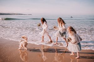 A group in white dresses splashes and plays together along a scenic beach at sunset with waves rolling in the background.
