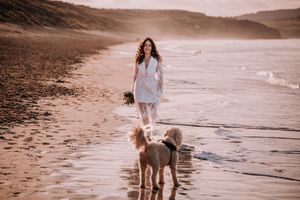 A person in a white dress walks along a misty beach shoreline with two dogs during golden hour.
